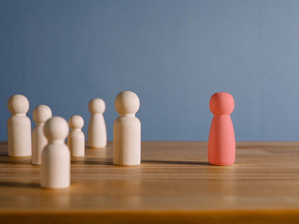 Red wooden figure standing alone, separated from a group of neutral figures, symbolizing emotional isolation and the need for psychological support or therapy - Katharina Treder psychotherapy online and in person